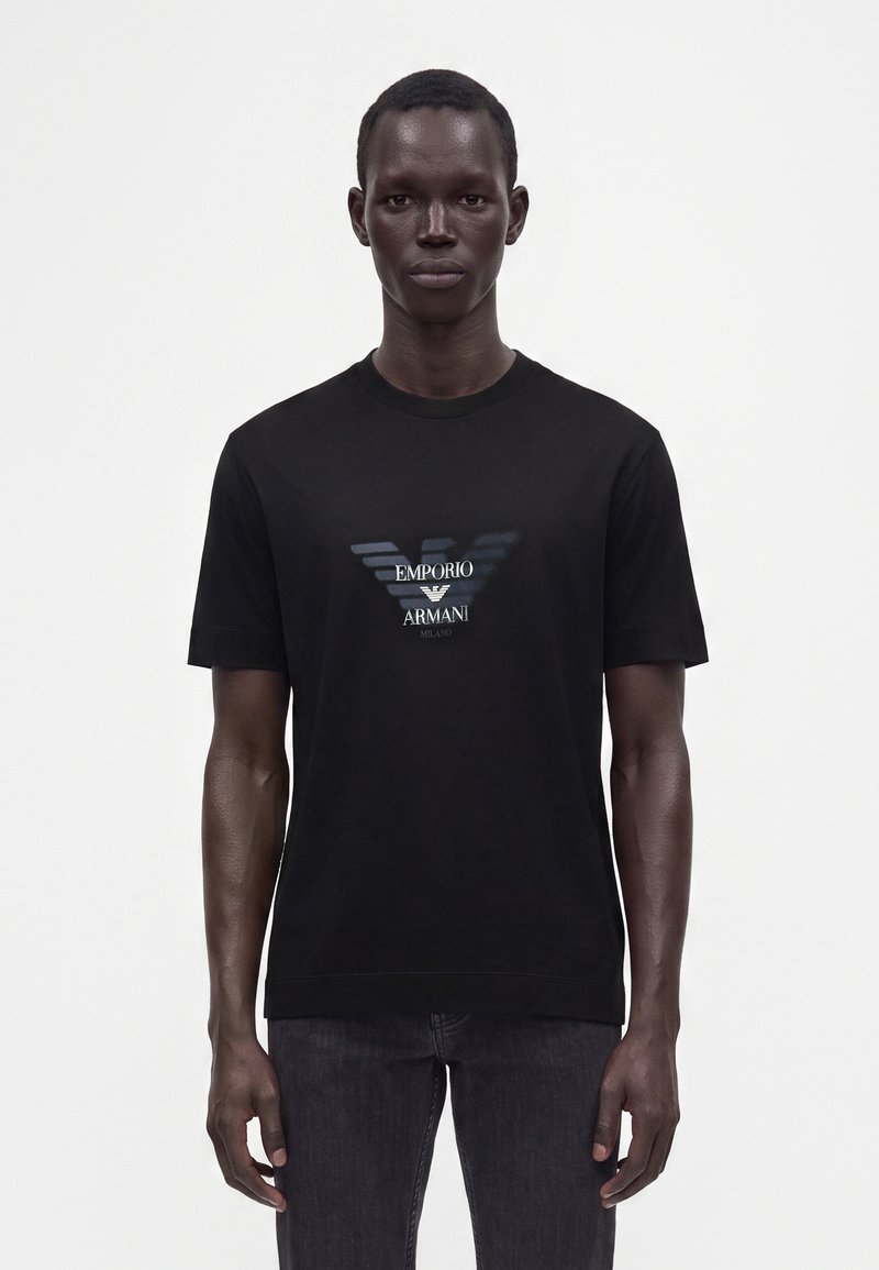 Άνδρας που φοράει μαύρο t-shirt Emporio Armani με εκτύπωση λογότυπου, όρθιος μπροστά σε απλό λευκό φόντο.