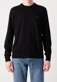 Homme portant un pull noir uni à manches longues associé à un jean denim bleu clair, debout devant un fond blanc uni.