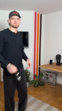 Zwart shirt met lange mouwen en broek, een bril vasthoudend. Op de achtergrond een houten tafel, plant, tv en een kleurrijke gestreepte muur.