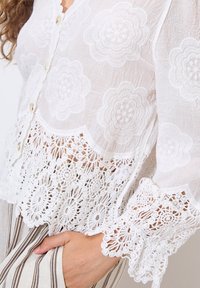 Weiße Bluse mit floraler Stickerei und Spitzenbesatz am Saum und an den Ärmeln. Mit Knöpfen vorne und strukturiertem Stoff.