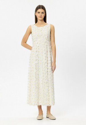 Frau mit langen braunen Haaren, die ein ärmelloses weißes Maxikleid mit einem Gänseblümchenmuster und beigefarbenen Ballerinas trägt, steht vor einem weißen Hintergrund.