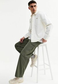 Witte lange mouwen shirt over een wit T-shirt, gecombineerd met olijfgroene cargobroeken. Beige sneakers met textuuraccenten.