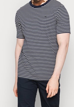 Homme portant un t-shirt à manches courtes rayé horizontalement bleu marine et blanc et un pantalon sombre, debout devant un fond uni.