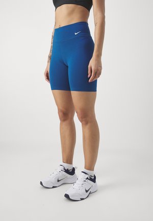 Femme portant un short de sport bleu taille haute, un soutien-gorge de sport noir, des baskets blanches Nike, posant devant un fond clair uni.