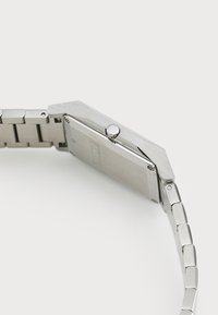 Reloj de acero inoxidable plateado con una caja rectangular, una superficie texturizada y una pulsera de eslabones. Presenta una corona circular en el lateral.