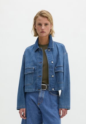 Blonde woman wearing a cropped blue denim jacket over an olive green top and high-waisted blue jeans with a black belt, facing forward.