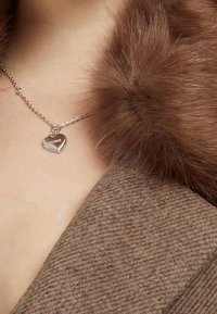 Collana a forma di cuore in argento con incanto "GUESS", abbinata a un colletto in finta pelliccia marrone, su un cappotto marrone dalle texture. 