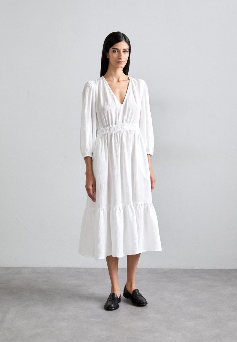 Robe blanche à manches longues avec un décolleté en V profond et une taille froncée, présentant une jupe superposée et un tissu texturé. Associée à des mocassins noirs.