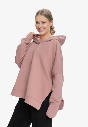 Młoda kobieta uśmiechająca się, ubrana w oversize'ową, zakurzoną różową bluzę z kapturem i czarne spodnie, trzymająca kołnierz bluzy jedną ręką.