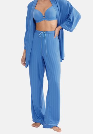 Kobieta ubrana w niebieski biustonosz, pasujące piżamowe spodnie ze sznurkiem oraz otwarty szlafrok, stojąca boso na białym tle.