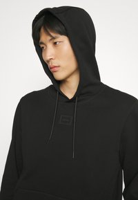 Sweat à capuche noir en tissu doux, avec une poche avant, des cordons de serrage, un patch logo sur la poitrine et une coupe décontractée avec une capuche.