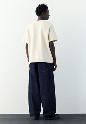 Oseba s kratkimi kodrastimi lasmi, oblečena v ohlapno bež majico in široke temno modre kavbojke, stoji obrnjena stran proti beli steni.