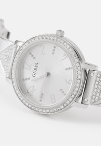 Sølv analog klokke med hvit urskive, krystallakkenter, timeindikatorer og et teksturert rem med innlagte rhinestones.