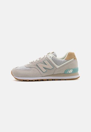 Lichtgrijze New Balance sneaker met beige accenten, turquoise logo en hielondersteuning, witte veters en gumzool, zijaanzicht op een egale achtergrond.