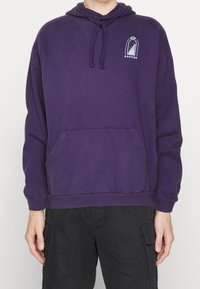 Lila bomullshuvtröja med en framficka, dragband kapuschong och en grafisk logotyp på bröstet. Har ribbade muddar och nederkant.