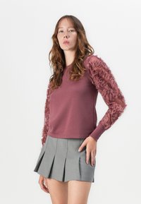 Maglione a collo alto in prugna con maniche trasparenti a volant, abbinato a una mini gonna grigia plissettata. Texture liscia con accenti morbidi in contrasto.
