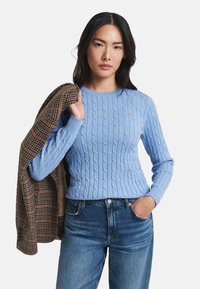 Lyseblå kabelstrikket genser med rund hals, kombinert med mørkeblå jeans. En rutete blazer henger over den ene armen.