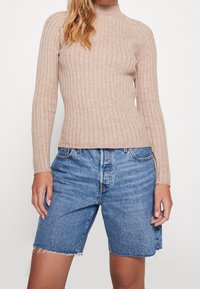 Top beige de punto acanalado con cuello alto falso, combinado con pantalones cortos de mezclilla azul con dobladillos deshilachados y silueta ajustada.