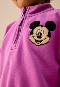 Fialová fleecová mikina na zip s texturovanou vyšitou nášivkou Mickey Mouse na levé části hrudi, s hladkým materiálem a stojáčkovým límcem.