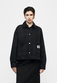 Άτομο που φοράει μαύρο σακάκι με κουμπιά και δύο μπροστινές τσέπες, η μία με λογότυπο Carhartt, σε συνδυασμό με μαύρη φούστα.