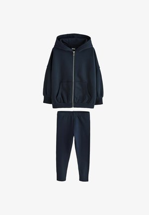 Ung pige med fletninger iført en mørkeblå hættejakke med lynlås, hvid skjorte, mørkeblå leggings og hvide sneakers, stående med det ene ben let bøjet.