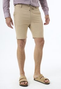 Shorts beiges en tissu texturé, avec une taille mi-haute et un cordon de serrage. Associés à des sandales en cuir marron clair à enfiler.