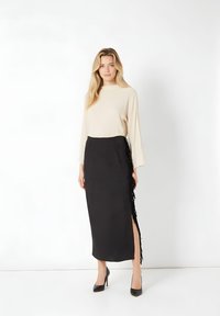 Mujer de pie con top beige de manga larga y falda midi negra con flecos laterales y abertura, usando zapatos de tacón alto negros, fondo blanco.
