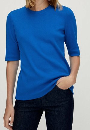 Persona che indossa una camicia a maniche corte di un blu acceso, infilata leggermente in jeans di denim scuro, con una mano in tasca.