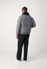 Doudoune grise avec un col noir, design matelassé, poches avant et poignets côtelés. Portée sur un pantalon noir et des baskets blanches.