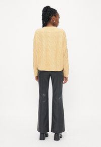 Pull en mailles torsadées jaunes, avec une coupe carrée, des poignets côtelés, associé à un pantalon en cuir noir évasé taille haute et des bottes à talons épais.