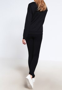 Mujer con pelo largo vestida con sudadera negra y leggins, alejándose, zapatillas blancas visibles sobre fondo claro.