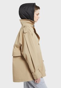 Beige puuvillatakki, jossa on suuri kaulus, musta huppu, nappisulku, sivutaskut ja käärityt hihat.