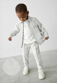 Harmaa teksturoitu verryttelypuku, jossa on vetoketjullinen takki, vastaavat housut ja valkoiset lenkkarit. Takissa on tasku, jossa on "BAKER"-merkki.