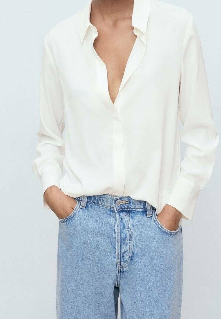 Personne portant une chemise blanche ample avec les boutons du haut déboutonnés et un jean taille haute bleu clair, les mains dans les poches.