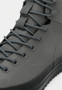 Gros plan d'une botte en cuir gris avec des œillets ronds, des lacets gris, une languette rembourrée et une semelle en caoutchouc noire.