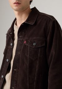 Bruine corduroy jas met een knoopsluiting aan de voorkant, voorzien van een kraag en twee borstzakken. Subtiele branding op de linkerzak.