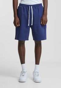 Navy blue cotton shorts with an elastic waistband and white drawstring. Features side pockets and a straight hem. Worn with white sneakers.