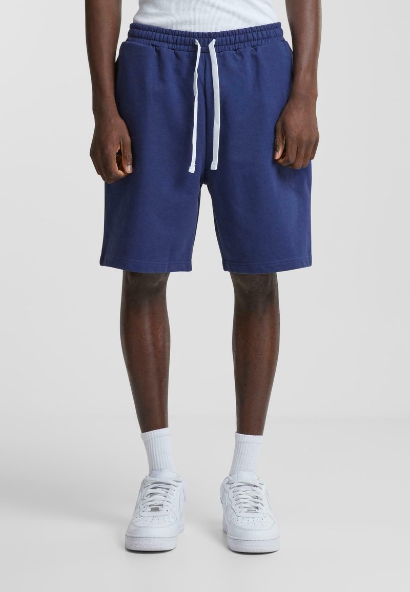 Navy blue cotton shorts with an elastic waistband and white drawstring. Features side pockets and a straight hem. Worn with white sneakers.