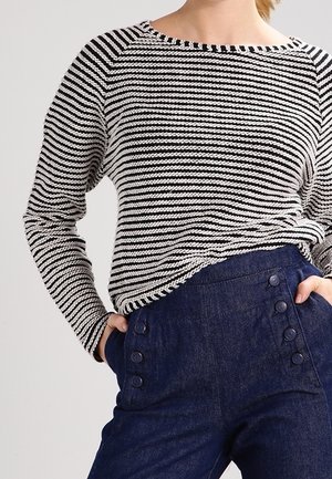 Osoba ubrana w czarno-biały top w paski z długim rękawem, wsunięty w wysokowalne, ciemnoniebieskie spodnie z sześcioma widocznymi guzikami.