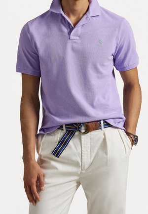 Mann trägt ein lilafarbenes Polohemd, cremefarbene Plisseehose mit einem blau-gelb gestreiften Gürtel und eine braune Lederuhr am linken Handgelenk.