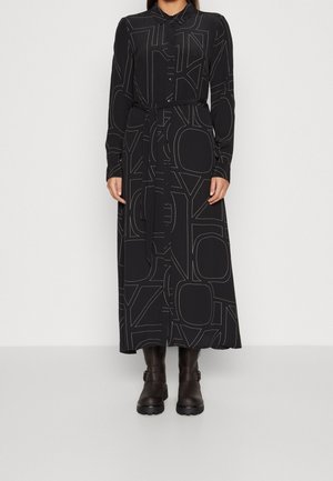 Robe noire longue avec un col, présentant un motif géométrique aux couleurs claires et une ceinture assortie, accompagnée de bottines noires.