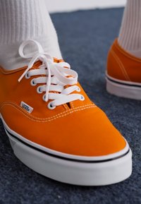 Narančasti platneni tenisice s bijelim vezicama, s crnim gumiranim potplatom i malim Vans logotipom sa strane, postavljene na strukturiranu pozadinu.