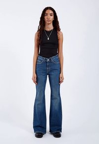 Laskostetut siniset farkut korkealla vyötäröllä, viidellä taskulla ja kevyellä kulutuksella. Yhdistetty istuvaan mustaan hihattomaan paitaan ja massiivisiin mustiin saappaisiin.