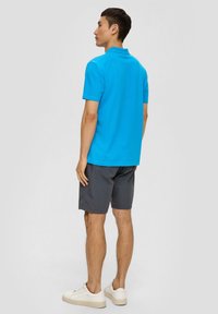 Hellblaues Polohemd mit Kragen, kurzen Ärmeln, kombiniert mit dunkelgrauen Shorts und weißen Sneakers, von hinten betrachtet.