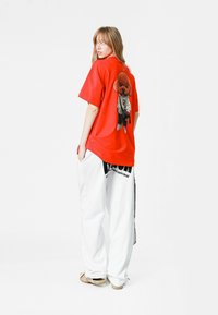 Camiseta roja de gran tamaño con un gráfico de un oso en la parte posterior. Combinada con pantalones blancos holgados y sandalias beige. Diseño simple, estilo casual.