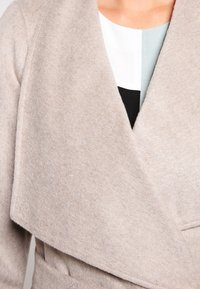 Cappotto avvolgente in misto lana beige con texture morbida, ampi rever e una vestibilità allentata. Design anteriore sovrapposto con finitura sartoriale.