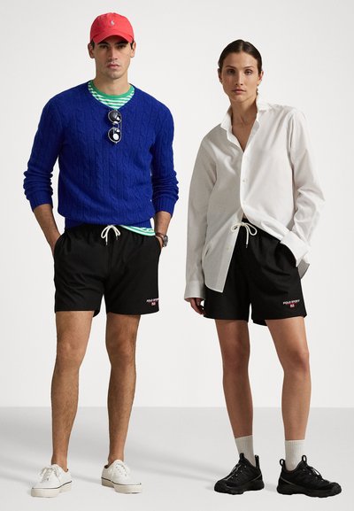 Blauer Kabelstrickpullover, weißes Hemd mit Knöpfen, schwarze Shorts mit Logo, rote Baseballkappe, schwarze Sneakers und weiße Slipper ausgestellt.