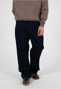 Mann trägt dunkelblaue, locker sitzende Jeans, braunes Sweatshirt und braune Mokassin-Lederschuhe und steht vor einem einfarbigen hellen Hintergrund.