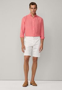 Camicia in lino rosa chiaro con bottoni e maniche lunghe, abbinata a pantaloni corti bianchi e mocassini beige, in piedi su uno sfondo grigio neutro.