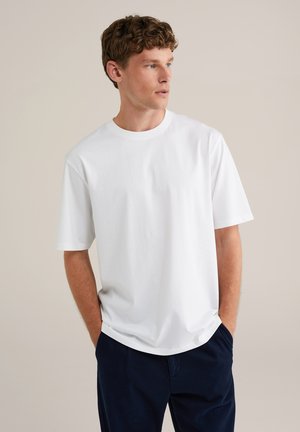 Camiseta de algodón blanca con mangas cortas, cuello redondo y corte holgado. Combinada con pantalones azul marino. Textura suave y diseño minimalista.